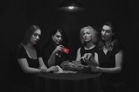 SZenenbild aus dem Kurzfilm 4/4. Vier Frauen mit rotem Lippenstift sitzen in Schwarz-Weiß-Optik an einem Tisch und spielen Karten, eine hält eine rote Spielkarte mit Logo in die Kamera.