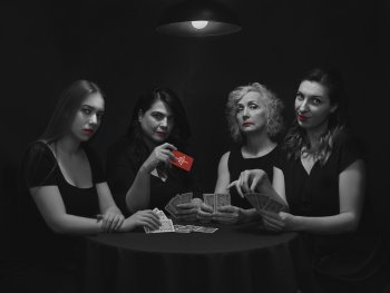 Copyright: Griechischer Kunstclub SZenenbild aus dem Kurzfilm 4/4. Vier Frauen mit rotem Lippenstift sitzen in Schwarz-Weiß-Optik an einem Tisch und spielen Karten, eine hält eine rote Spielkarte mit Logo in die Kamera.