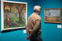 Eine Person mit grauem Haar und Brille geht in einer Ausstellung an zwei Gemälden vorbei, die an einer türkisfarbenen Wand hängen: links eine Szene mit einer liegenden Person und einer Figur an einem Baum, rechts eine Birkenlandschaft.