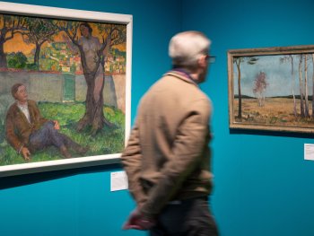 Eine Person mit grauem Haar und Brille geht in einer Ausstellung an zwei Gemälden vorbei, die an einer türkisfarbenen Wand hängen: links eine Szene mit einer liegenden Person und einer Figur an einem Baum, rechts eine Birkenlandschaft.