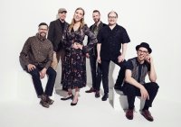 Sechsköpfige Musikgruppe in stilvoller Kleidung posiert lächelnd vor weißem Hintergrund.