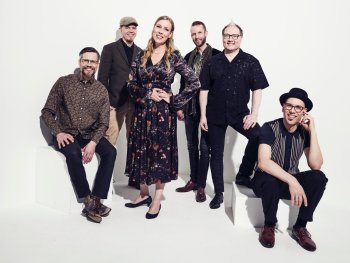 Sechsköpfige Musikgruppe in stilvoller Kleidung posiert lächelnd vor weißem Hintergrund.