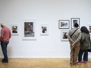 Drei Besucherinnen und Besucher betrachten in einer Galerie gerahmte Farbfotografien von Menschen im Straßenumfeld, darunter Porträts mit Kofferradios und Motorrädern.
