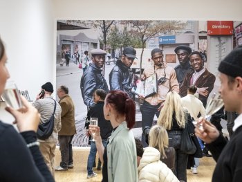 Besucherinnen und Besucher einer Ausstellungseröffnung stehen mit Sektgläsern vor einem wandgroßen Foto, das fünf Männer in einer Einkaufsstraße zeigt.
