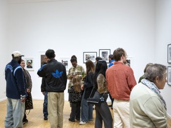 Viele Personen stehen und unterhalten sich in einem hellen Ausstellungsraum mit gerahmten Fotografien an den weißen Wänden.