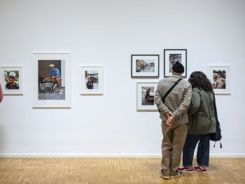 Zwei Besucher betrachten in einer Galerie gerahmte Farbfotografien an einer weißen Wand, die Porträts junger Menschen mit Kofferradios und Motorrädern auf der Straße zeigen.
