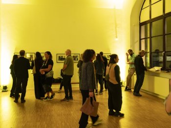 Besucherinnen und Besucher stehen in einem gelb beleuchteten Ausstellungsraum, unterhalten sich und betrachten gerahmte Schwarz-Weiß-Fotografien an den Wänden.
