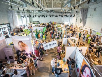 Gut besuchte Messe in einer hohen Industriehalle mit vielen Ständen für vegane und nachhaltige Produkte, zwischen denen Besucherinnen und Besucher entlangschlendern.