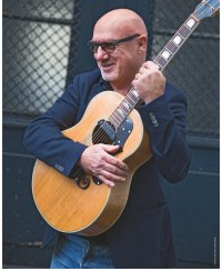 Lächelnde Person mit Glatze, Brille und dunklem Sakko hält eine Akustikgitarre mit hellem Korpus fest im Arm und steht vor einem dunklen Gittertor.