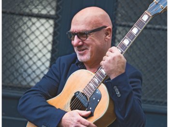 Copyright: Achim Weinberg Lächelnde Person mit Glatze, Brille und dunklem Sakko hält eine Akustikgitarre mit hellem Korpus fest im Arm und steht vor einem dunklen Gittertor.