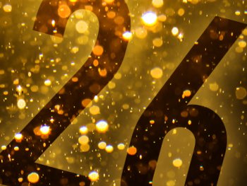 Goldene, glitzernde Lichtpartikel auf dunklem Hintergrund mit der groß gestalteten Zahl „26“, die an eine Jahreszahl zum Jahreswechsel erinnert.