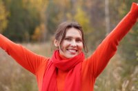 Lächelnde Person in orangefarbenem Pullover und rotem Schal streckt im herbstlichen Wald die Arme in die Höhe.