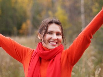 Lächelnde Person in orangefarbenem Pullover und rotem Schal streckt im herbstlichen Wald die Arme in die Höhe.