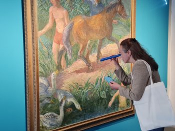 Eine Person mit Stofftasche betrachtet in einer Ausstellung durch eine blaue Rolle ein großes Gemälde mit Pferd, Gänsen und badenden Menschen.