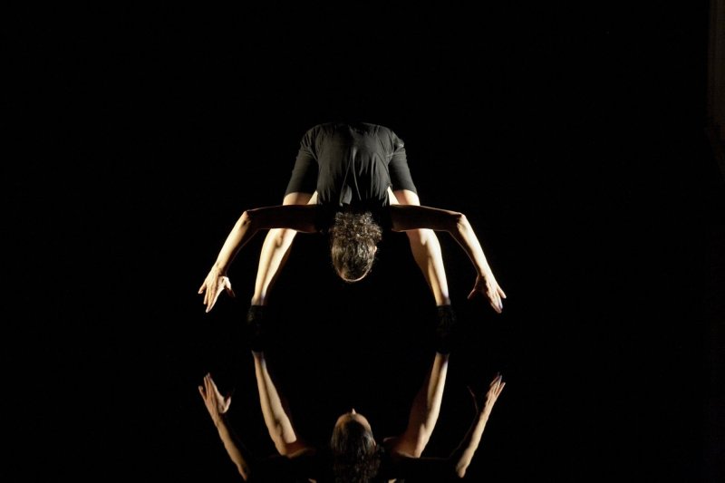 A woman stands with her legs apart in front of a black background. Her upper body is bent far towards the floor and she holds her arms at right angles towards the floor. Her reflection can be seen on the floor.