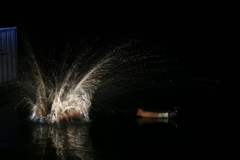 Eine Person fällt vor schwarzem Hintergrund auf den mit Wasser bedeckten Boden, wodurch ein großer Wasserspritzer entseht.