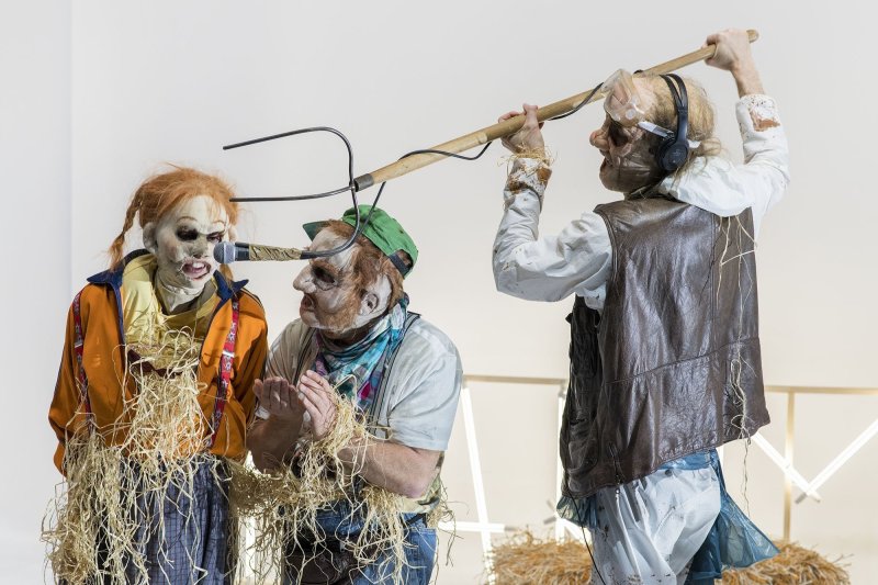 Three people dressed as scarecrows. The person on the far right is holding a pitchfork with a microphone to the person on the far left who is speaking into it.