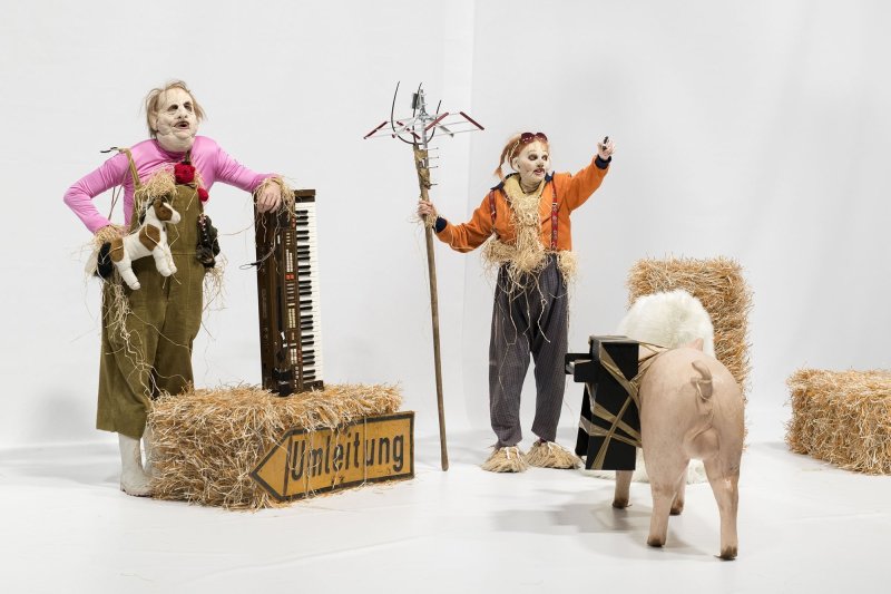 Two people dressed as scarecrows in front of a white background. One leans against a vertically positioned keyboard, the other holds a pitchfork with pieces of metal. Hay bales and a dummy pig are placed around them.