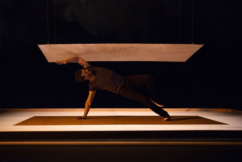A man leaning sideways against the ground. He is supporting himself with one hand, while the other is stretched towards the wooden board, that‘s hovering closely above him.
