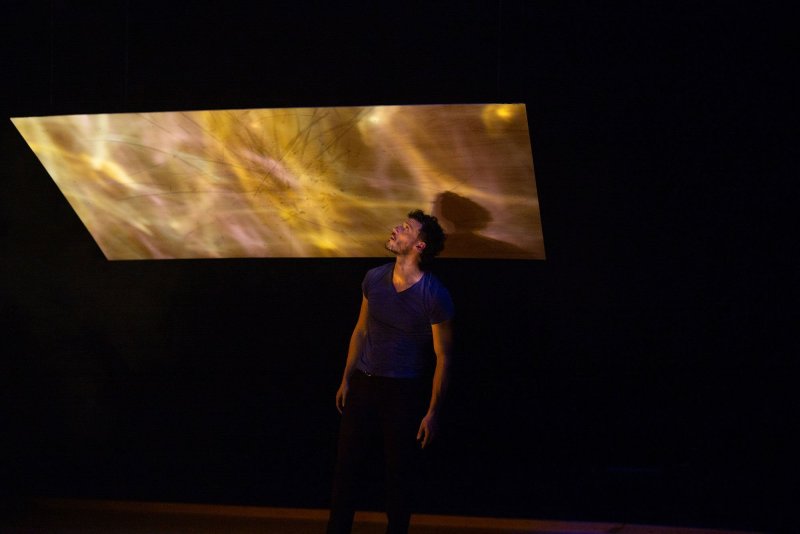 A man on a dark stage. Directly above him floats a wooden board, which is a little taller and wider than himself.