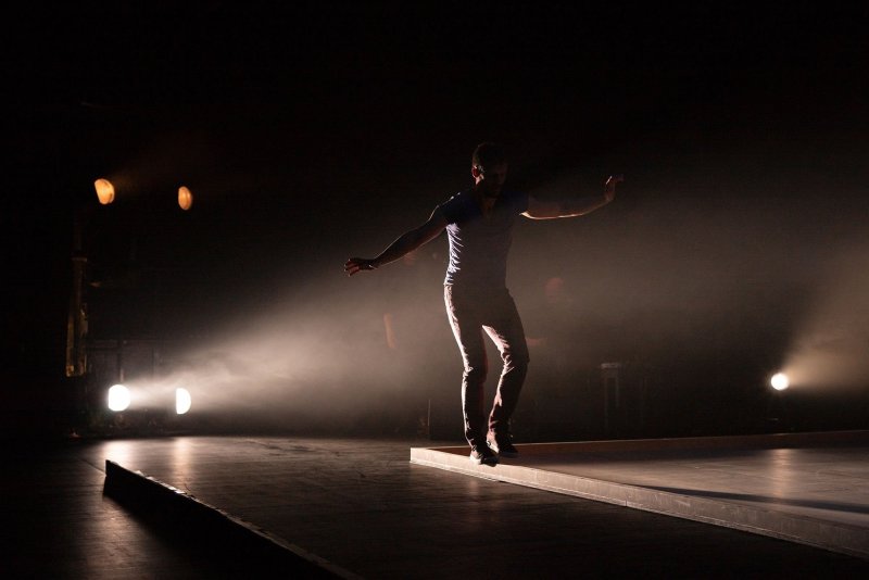 A man on a dark stage, only being lit from behind. He’s balancing on a black wodden frame.