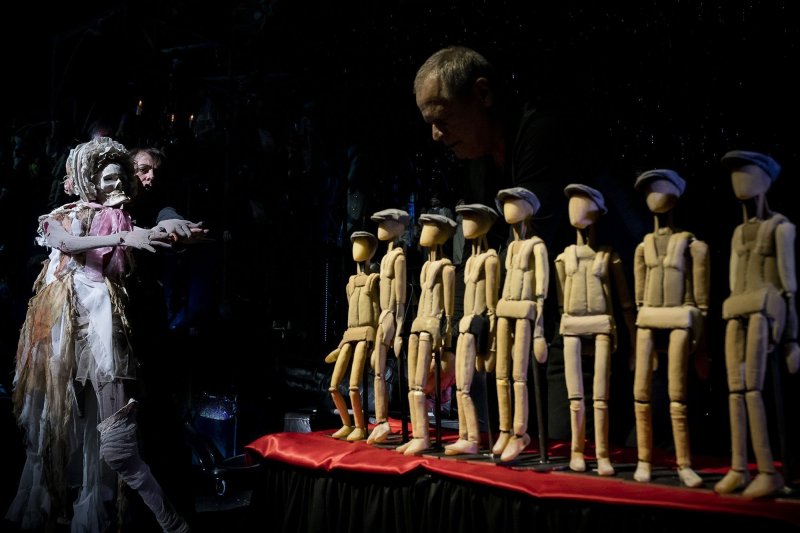 Several puppets with slate hats are lined up on a table covered with red fabric. Behind them stands a man disappearing into the shadows. A man enters the picture from the left, controlling a ghost-like, human-sized hand puppet.