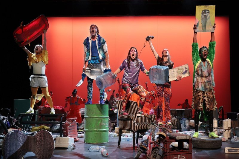 Five people can be seen on a heavily cluttered stage. They are standing on various pieces of furniture and objects, holding up all kinds of objects and shouting. Behind them are several people in orange high-visibility gear.