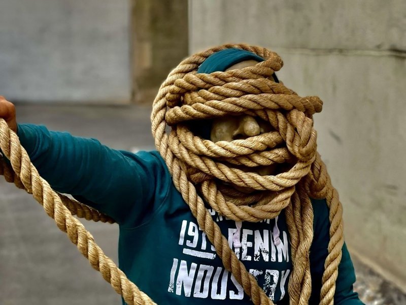 A man wearing a face mask and green hooded sweatshirt, whose face is almost entirely wrapped in thick rope.