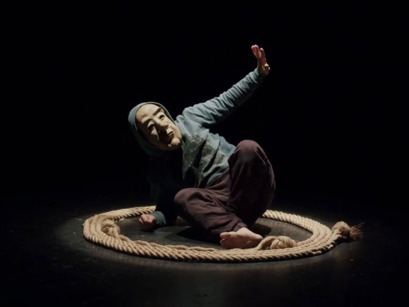 A man wearing a mask and a green hoodie is lying on the ground in a hunched posture. He is stretching his left arm in the air while looking after it. He is surrounded by a thick rope lying on the ground.