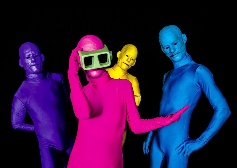 Vier Personen in farbigen Ganzkörperanzügen und Masken über den ganzen Kopf posieren. Die Person im Vordergrund trägt eine übergroße gebastelte VR-Brille.