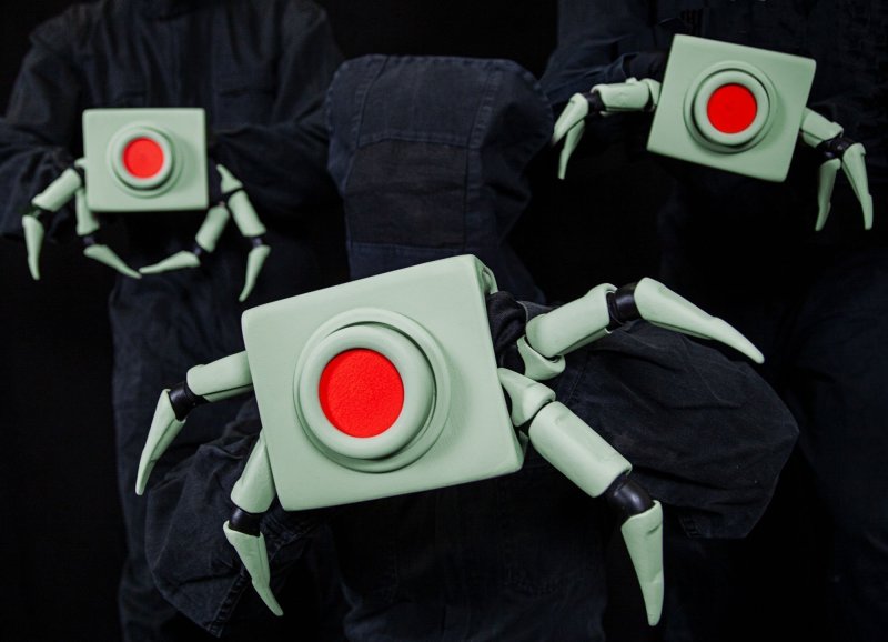 Close up of three crab-like robots with a big red button. They hang on the backs of three hooded people.