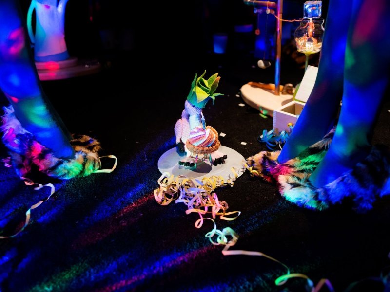 Close up of a small stuffed animal sitting in front of a fabric birthday cake. Two people in fur-covered high-heeled shoes are standing around him, the scenery is bathed in disco lights.