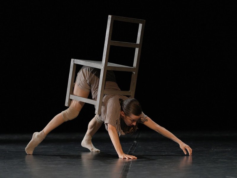 A dancer on stage. She is walking on all fours. She carries a wooden chair on her body, similar to a snail shell.