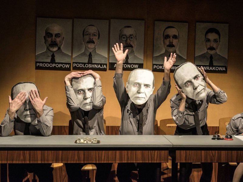 Vier Personen sitzen in einer Reihe an einem Holztisch. Sie tragen alle graue Kleidung und eine große Maske aus Pappe, während jeder auf eine andere Weise gestikuliert.