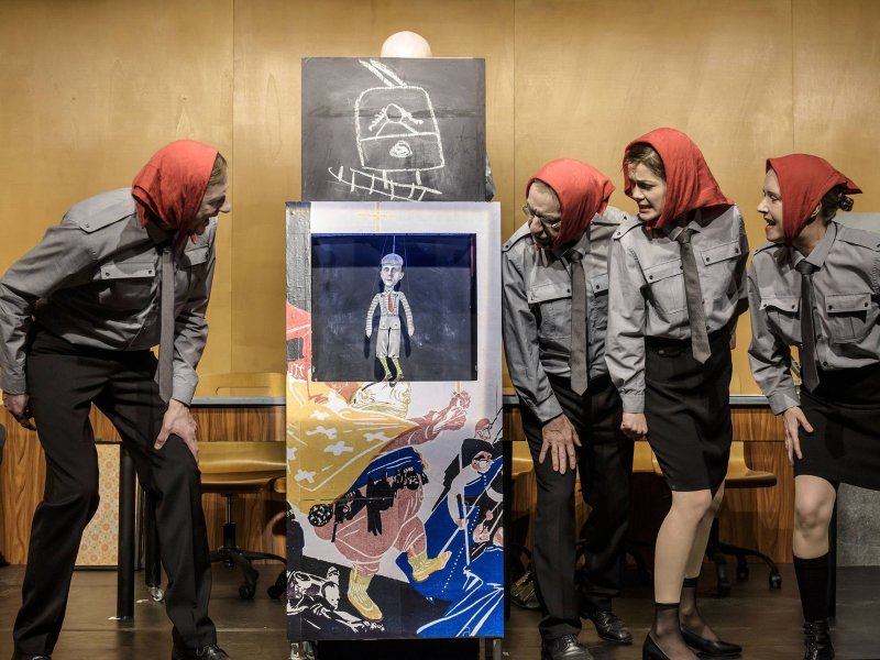 Four people in red headscarves and gray shirts look intently at a small paper puppet of a man hanging between them.