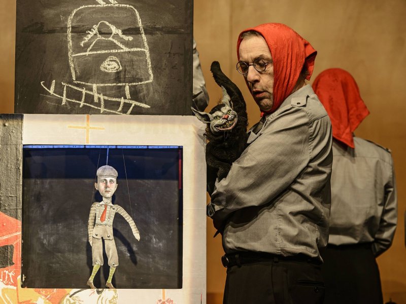 A person with a red headscarf, round glasses and gray shirt stands in the foreground of the picture holding a mean looking cat puppet. Next to him is a blackboard with a train drawn onto it, and a paper puppet in a wooden frame.