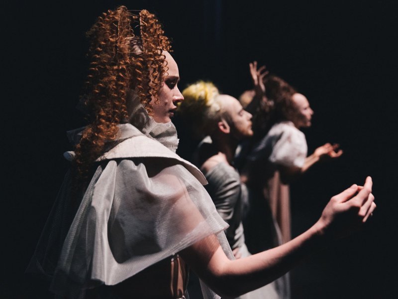 Three people with gray, translucent clothes and curly hair stand lined up on a dark stage.