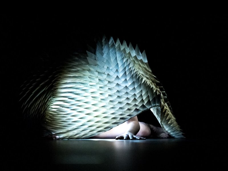 A person's arms and legs peek out from under a white sculpture, made out of hundreds of paper cones, which is lilluminated from the inside.