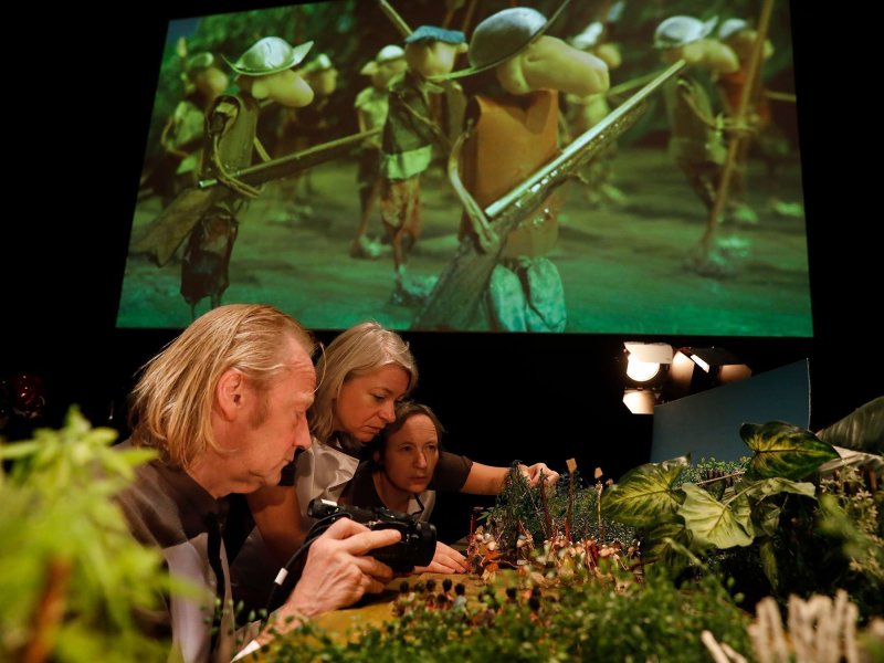 Shot of a table with a handcrafted landscape with small human figures controlled and filmed by three real people. The game scenery is projected onto a screen in the background.