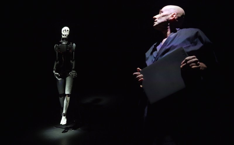 Ein Mann in schwarzer Kleidung steht im Vordergrund einer dunklen Bühne. Er sieht einen hinter sich stehenden, weißen Roboter an.