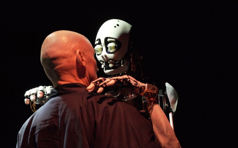 A bald man is shown from behind. In front of him, there is a white robot of about the same height, which has its arms placed on the man's shoulders.
