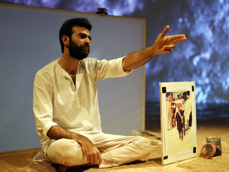 A man in white clothing is sitting cross-legged on the ground, which is covered in wheat seed. He is stretching his arm forward and forms a weapon with his hand. In front of him a picture of a child and a man is placed, as well as a jar filled with earth.