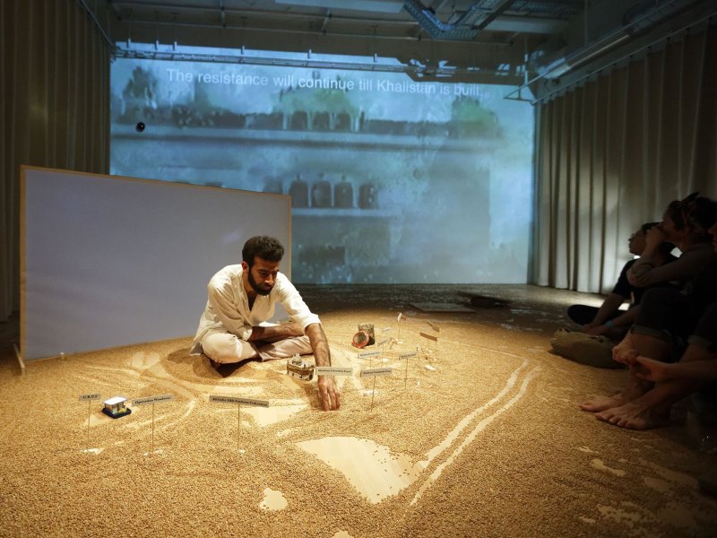 : A man in white clothing is sitting cross-legged on the floor, which is covered in wheat seed. In front of him are two small model houses and a jar of soil. Several small signs with various captions are placed in front of him. Behind him, a house with the caption 