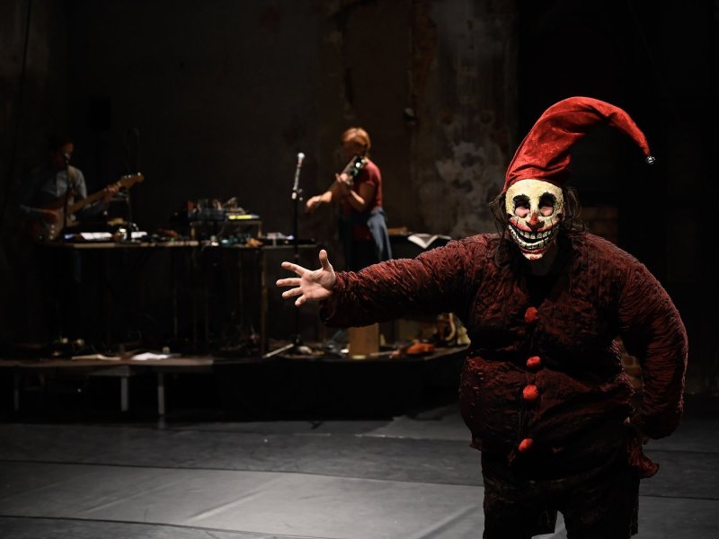 Eine Person mit roter Jacke, Zipfelmütze und grinsender Maske, die ihren rechten Arm nach vorne streckt, steht im rechten Vordergrund des Bildes.  Hinter ihr sind zwei Leute zu sehen, die Instrumente spielen.
