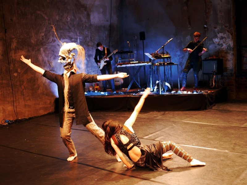 A woman sits on the floor and supports herself with her left arm, while stretching her right arm into the air. Next to her stands a person who is stretching both arms away from his body while looking upward. A large skellet mask with head and beard hair hovers where its head is meant to be. Behind them a band is playing several instruments.