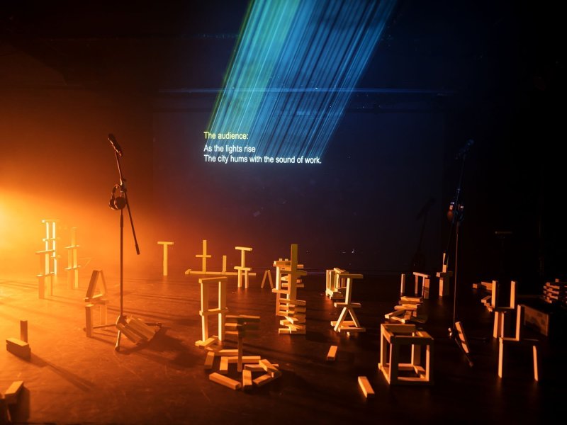 A dark stage, with a bright orange light shining onto it from the left border of the picture. A microphone, including a stand with a pair of headphones hanging from it, as well as various structures built from yellow building blocks are placed on stage. A projection in the background says: „The audience: As lights rise/ The city hums with the sound of work“