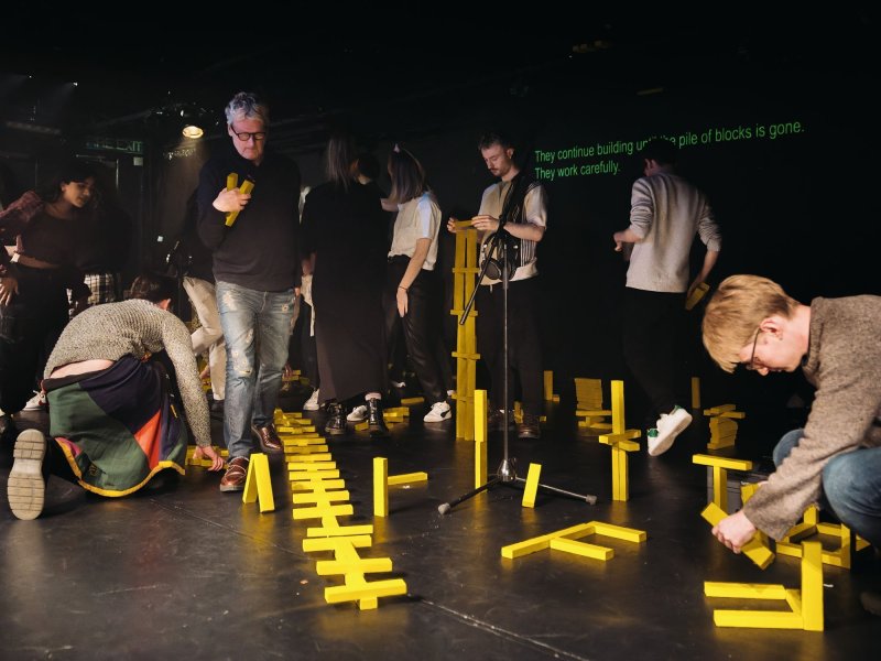 Several people build constructions from yellow building blocks on a black stage.