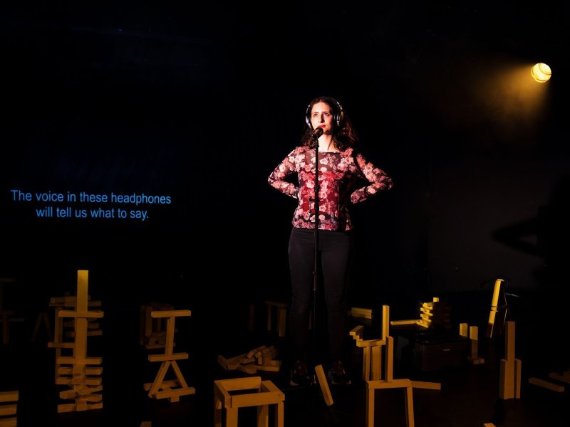 A woman wearing headphones on a dark stage with her hands on her hips. In front of her is a microphone, and around her are several constructions made of yellow building blocks. Behind her the text 