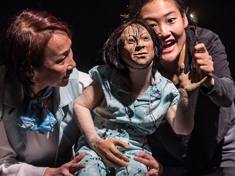 Close-up of two women controlling a human doll while looking at it very intensely. One of the two women is holding a spoon to the puppet’s hand.
