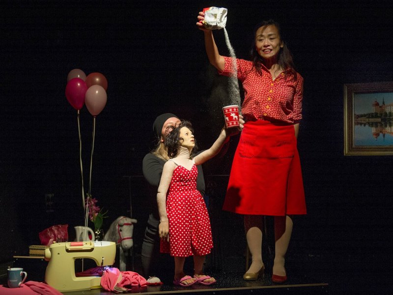Photograph of a stage with an actress in a red dress pouring sugar into a cup held by a hand puppet. The hand puppet looks like the actress and is controlled by a person standing behind her.